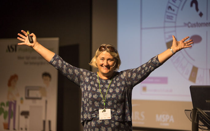 Alison stands in the centre of the picture with both arms raised, a smile on her face stood in front of a projected screen