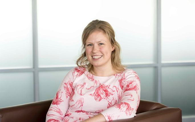 Nicola Beesley sits in the centre of the photo on a brown leather armchair smiling into the camera wearing a pink and white long sleeved top