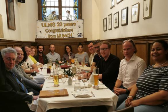 Alumni group posing for a photo around a table at a restaurant with a sign 