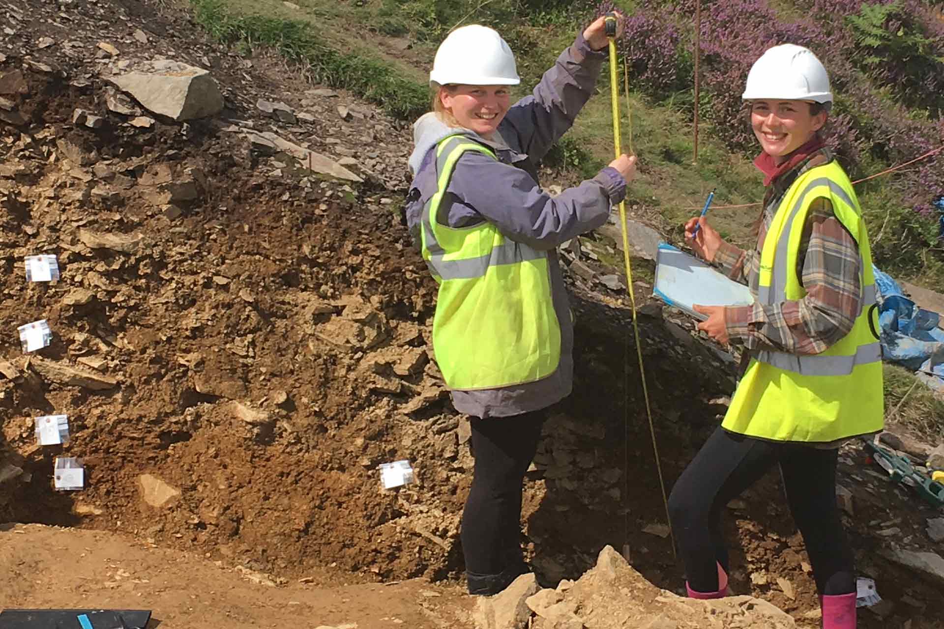 Two students on an archaeological site wearing hardhats and fluorescent jackets, one holding a tape measure and the other holding a clipboard.