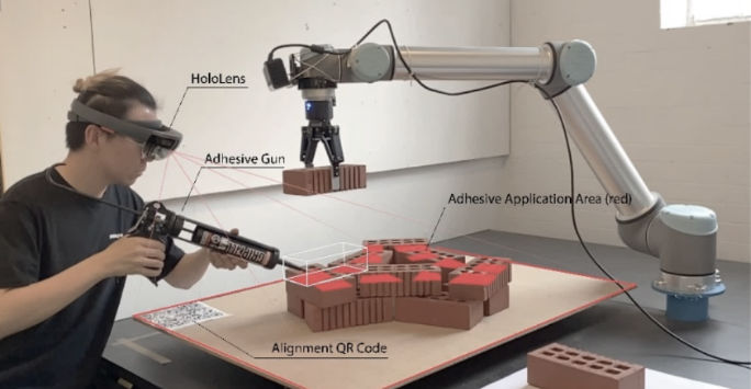 A man wearing virtual reality goggles holds an adhesive gun over a brick structure whist a robot arms holds a brick in place ready to be glued.