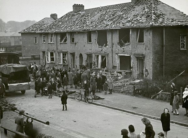 Black and white photo of a crowd standing next to bomb damaged houses.