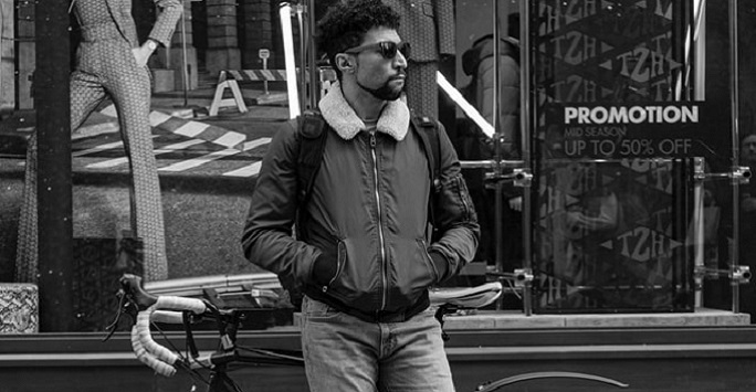 black and white photo of a young man in sunglasses standing in front of a shop front and bicycle