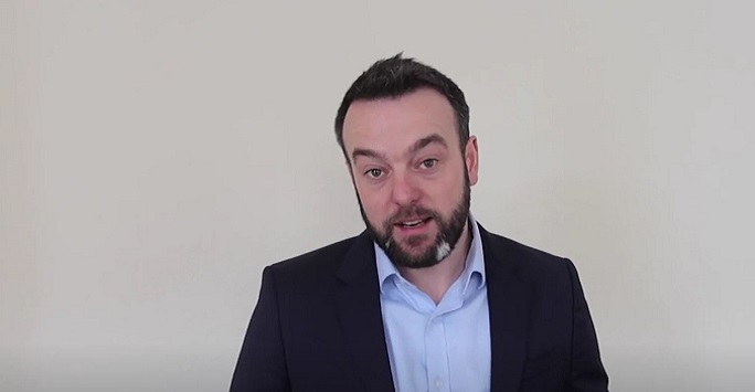 sldp leader colum eastwood in a blue shirt and black jacket standing in front of a white wall, talking to camera