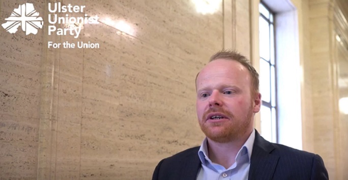 UUP member John Stewart in a light blue shirt with black jacket, interviewed in front of a light stone wall