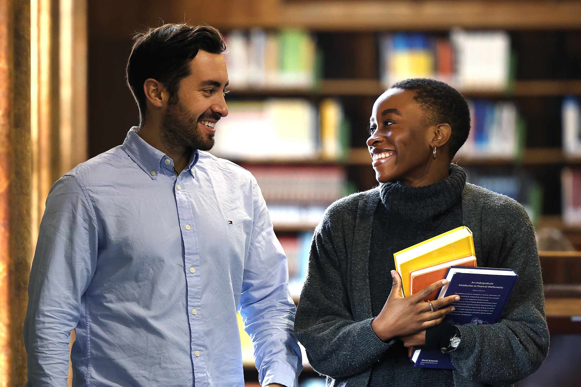 Two students walk through the library, one carrying some books.