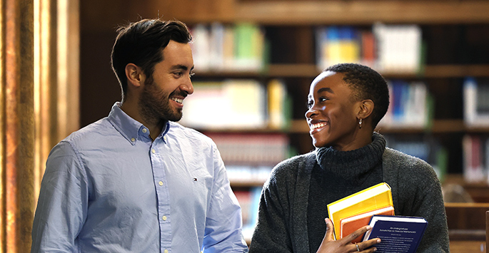 Two students walking in a library, one carrying some books.