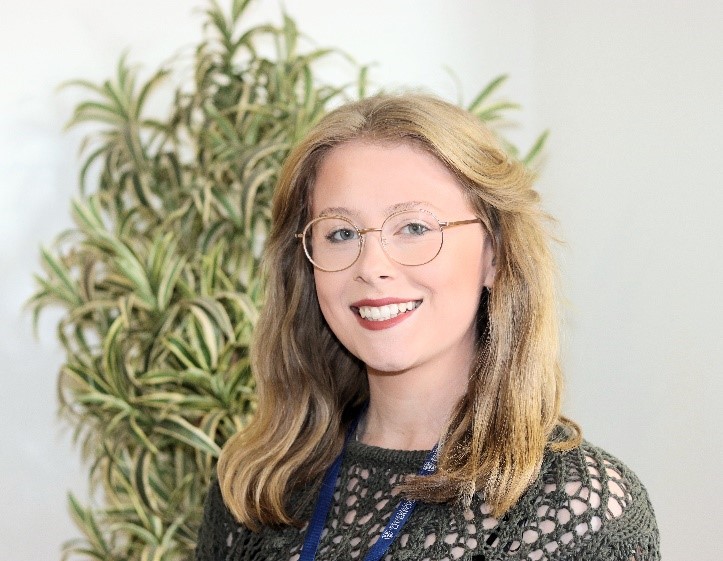 A picture of a white woman with shoulder length blonde hair wearing circular glasses and a green crocheted top, with a variegated spider plant in the background