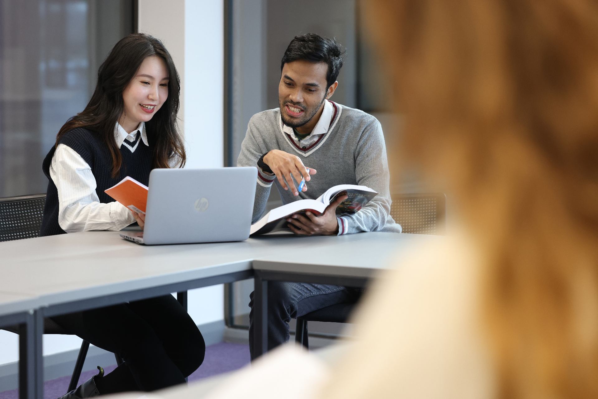 Two students sitting together at a desk, working on a laptop and with a text book.